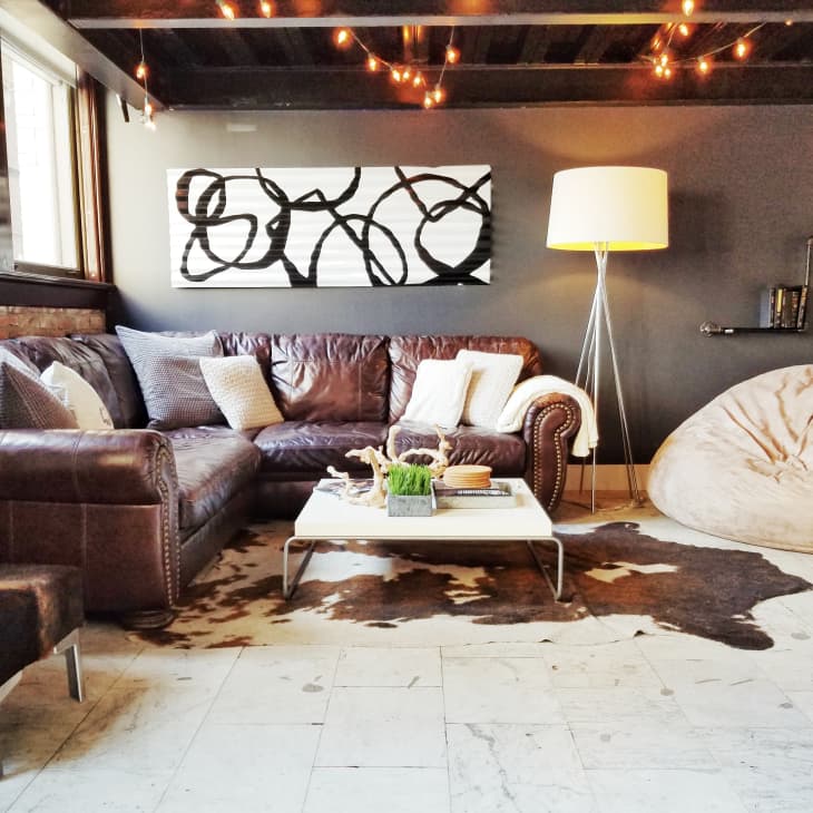 Cozy living room with brown leather sectional, abstract art, floor lamp, bean bag, and cowhide rug under string lights.