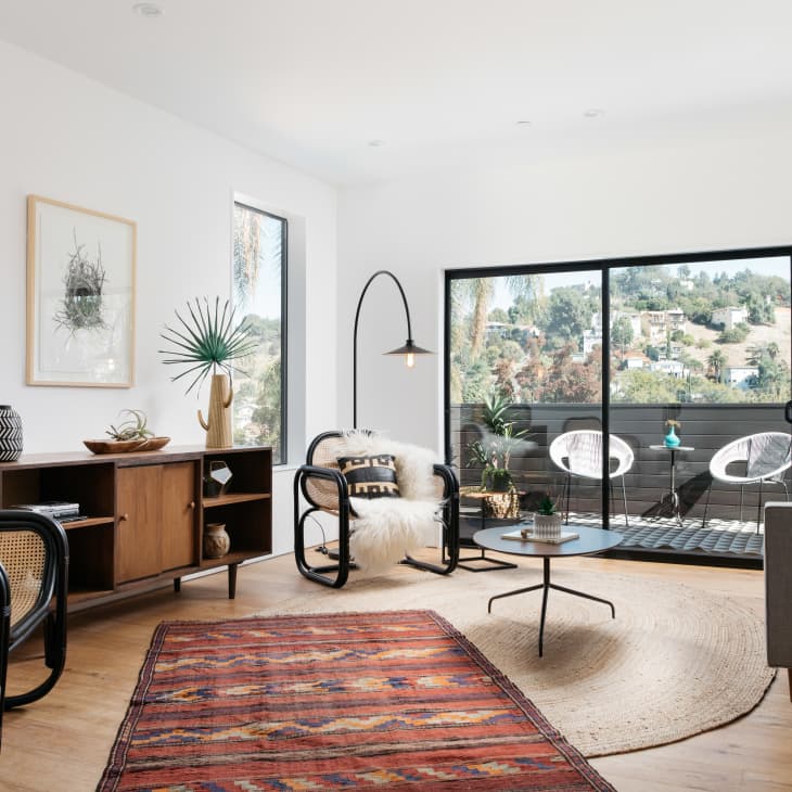 Modern living room with large windows, mid-century furniture, plants, and a colorful rug. Outdoor view with hills.