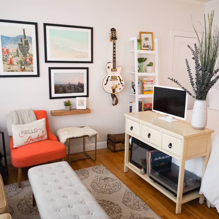 Cozy bedroom with a bed, orange chair, wall art, guitar, TV on a console, and bookshelf with decor and books.