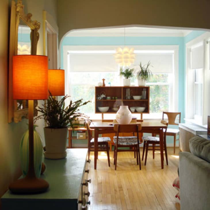 Dining room with wooden table, chairs, pendant light, and plants on a shelf by large windows.