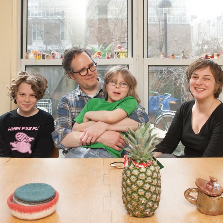 Family of four sitting at a table with a pineapple, teapot, and coasters, smiling in front of large windows.