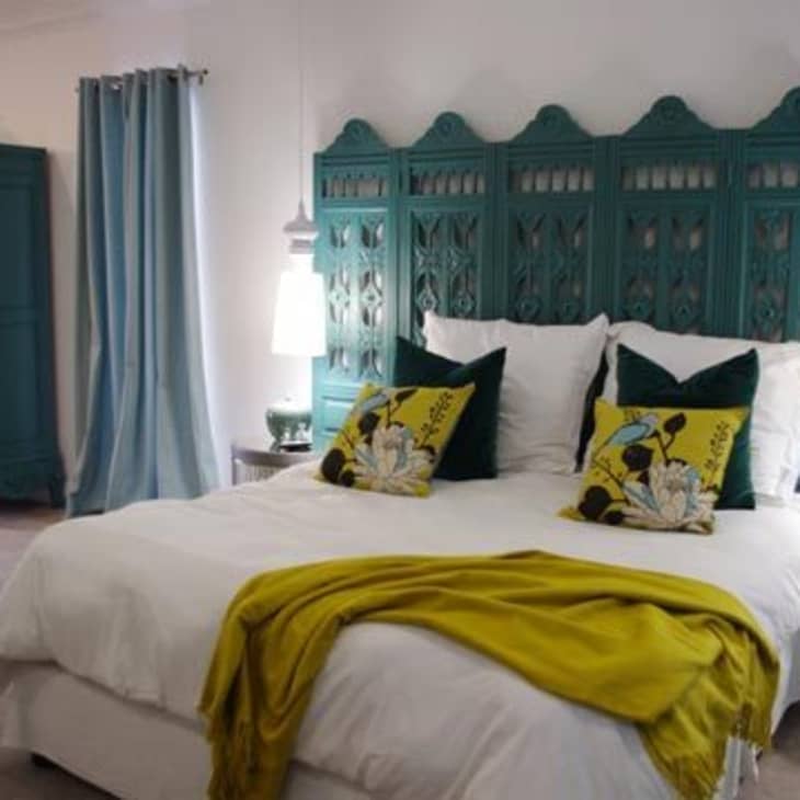 Bedroom with teal carved headboard, white bedding, yellow throw, and decorative pillows. Teal wardrobe and mannequin in the corner.