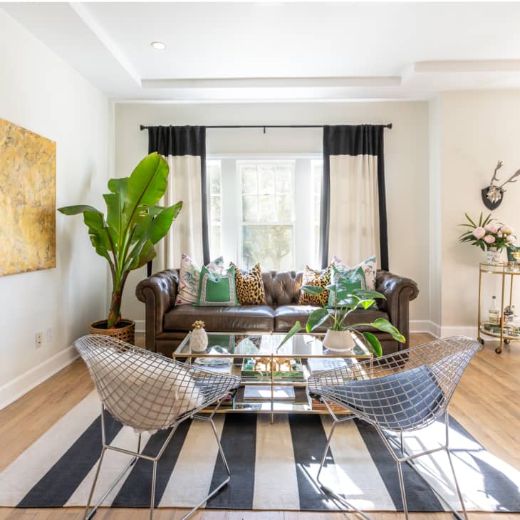 Living room with brown leather sofa, patterned cushions, glass coffee table, metal chairs, large plant, and abstract wall art.
