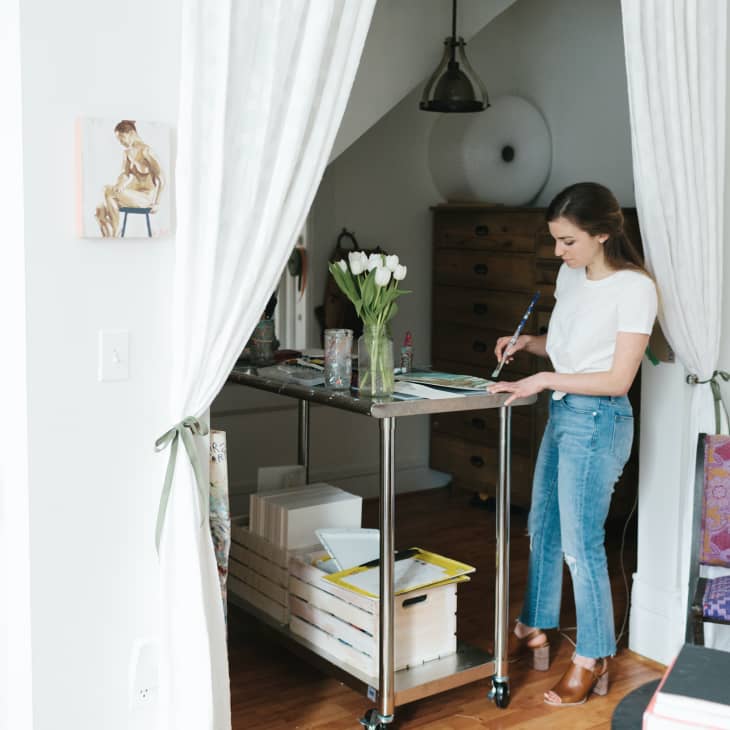 Woman painting at a rolling cart in a cozy room with white curtains, framed art, a ukulele on the wall, and a purple chair.