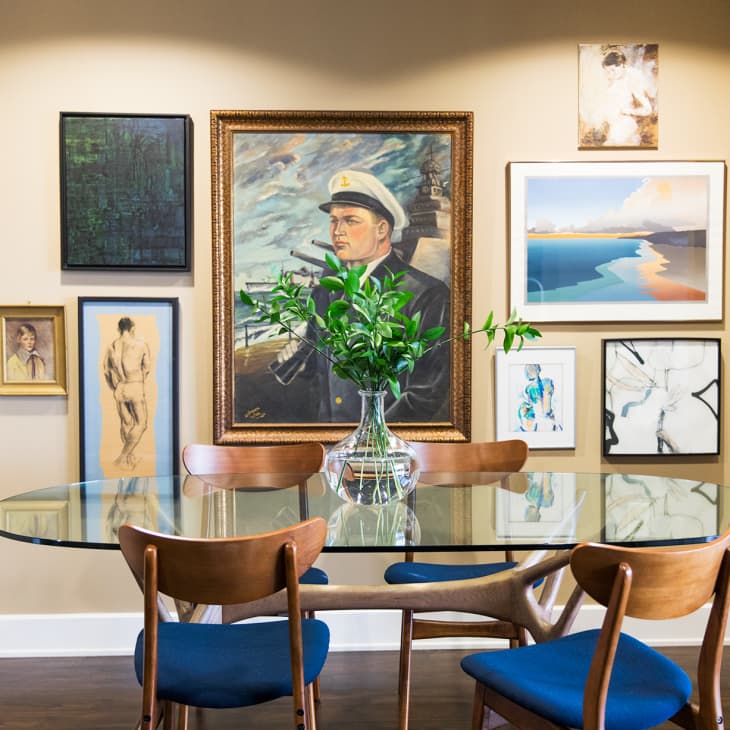 Glass dining table with wooden chairs, blue cushions, and a vase of greenery, surrounded by eclectic wall art.