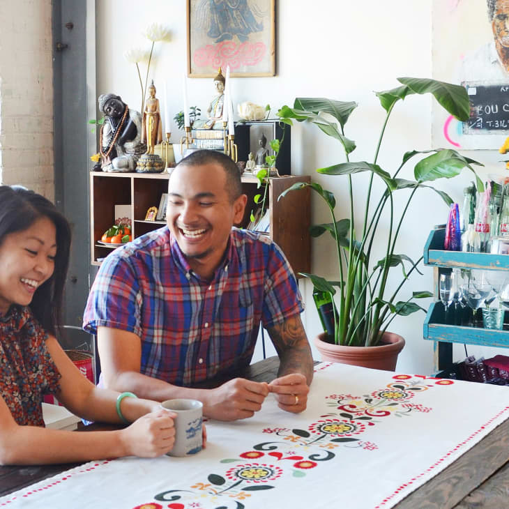 Two people smiling at a table with a colorful tablecloth, surrounded by plants and eclectic decor.