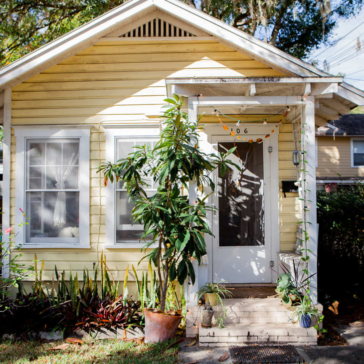 Yellow cottage with white trim, potted plants on porch, and string lights above door.
