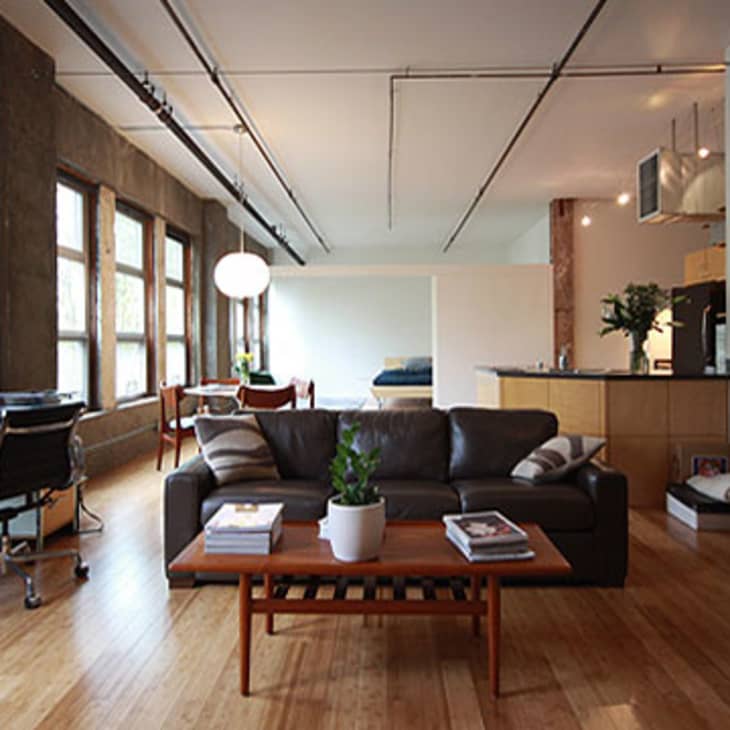 Open-plan living room with brown leather sofa, wooden coffee table, desk, bookshelves, and kitchen in the background.