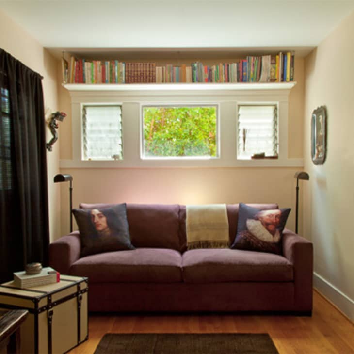 Cozy living room with a brown sofa, decorative pillows, bookshelves above, and a window view of greenery.