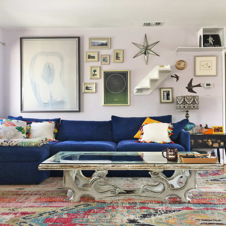 Living room with blue sectional sofa, colorful pillows, eclectic wall art, ornate glass coffee table, and vibrant rug.