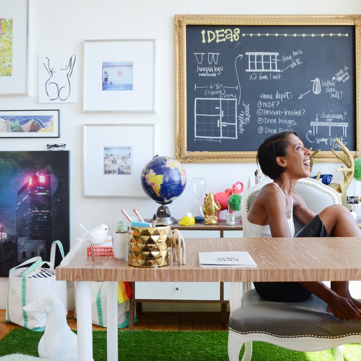 Woman sitting at a desk in a colorful, art-filled room with a chalkboard, globe, and eclectic decor.