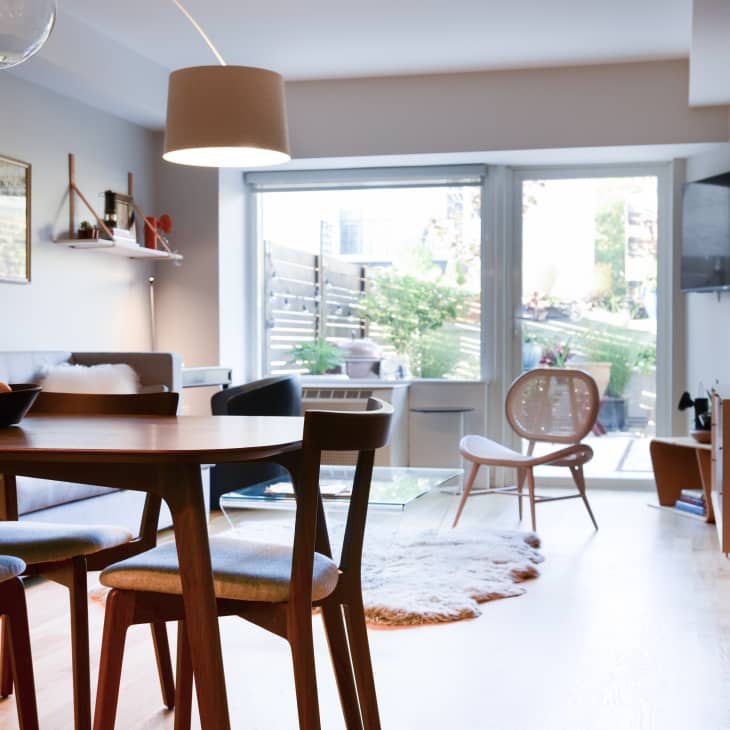 Modern living room with wooden dining table, blue chairs, wall art, and large window overlooking a garden.