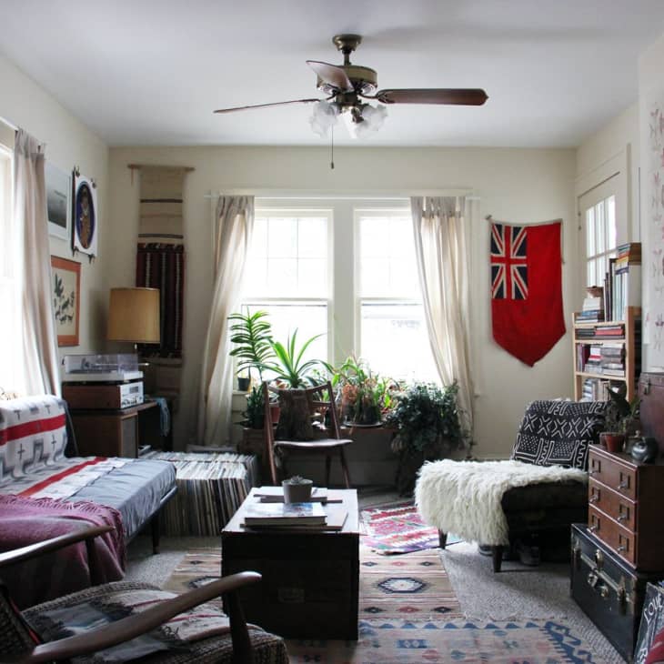 Cozy living room with eclectic decor, plants, bookshelves, vintage furniture, and a red flag on the wall.