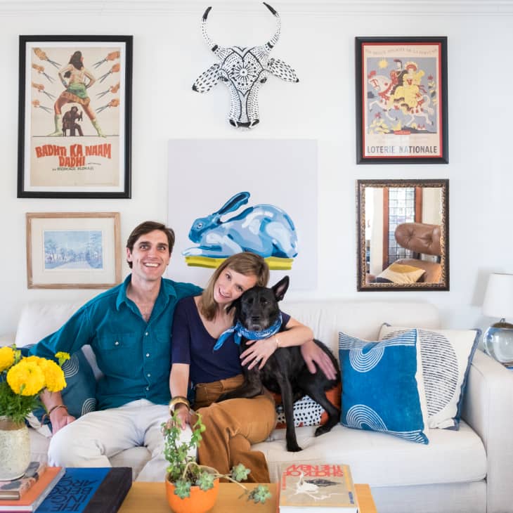 Couple sitting on a white sofa with a black dog, surrounded by colorful art, bookshelves, and yellow flowers in a vase.