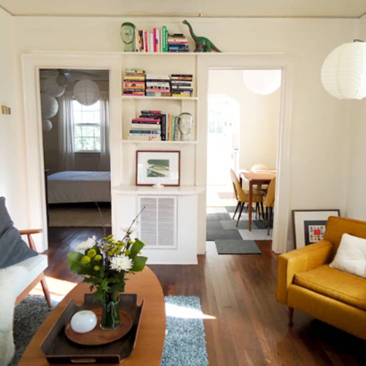 Living room with mustard armchair, blue sofa, bookshelf, and coffee table with flowers, leading to dining area and bedroom.