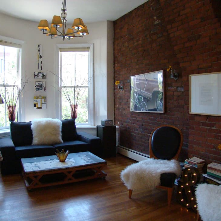 Living room with exposed brick wall, black sofa, fluffy white pillows, wooden floor, and modern chandelier.