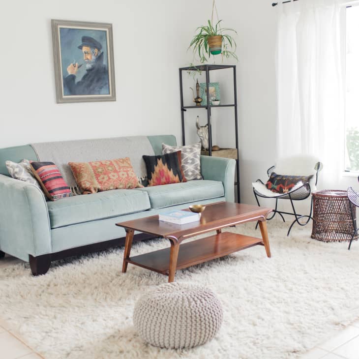 Living room with a light blue sofa, colorful cushions, wooden coffee table, and a plant on a shelf by a large window.