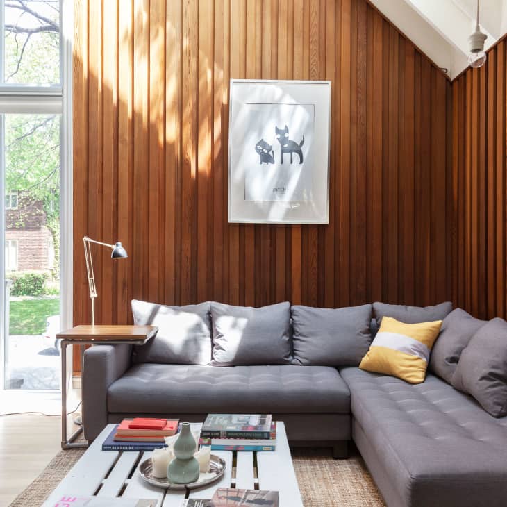 Gray sectional sofa with yellow pillow in a living room with wood panel walls, large window, and framed cat artwork.