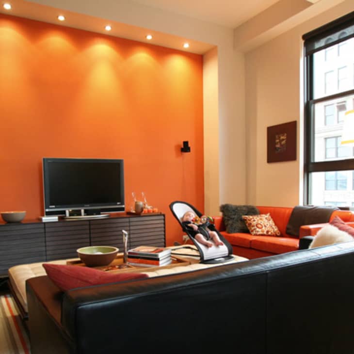 Modern living room with orange accent wall, black TV stand, red sectional sofa, and large window with city view.