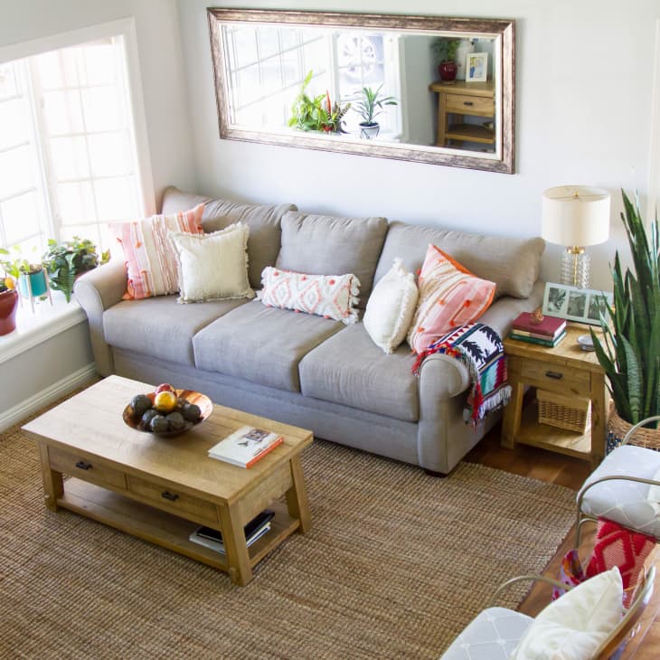 Cozy living room with a gray sofa, colorful pillows, wooden coffee table, large mirror, and potted plants by the window.