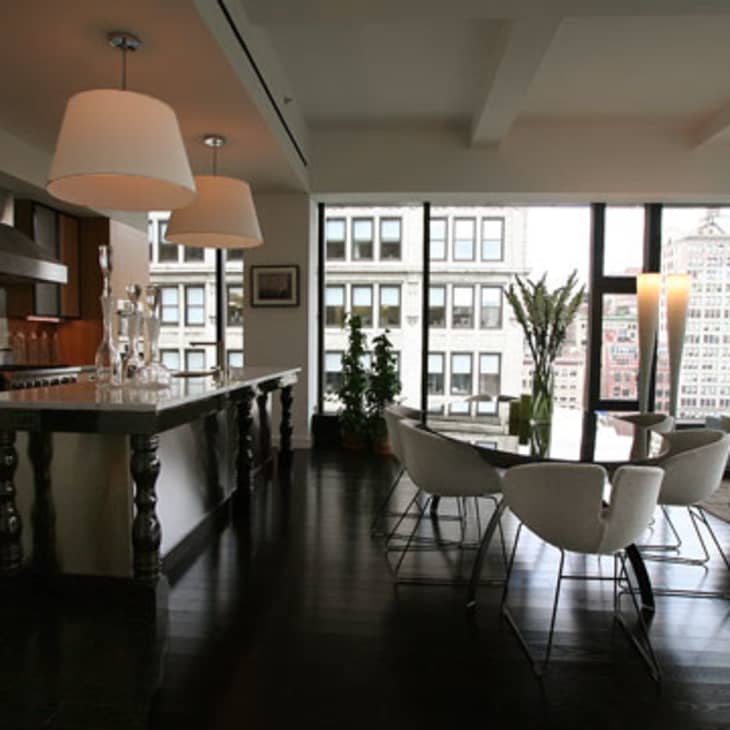 Modern open-plan kitchen and living area with dark wood floors, large windows, white chairs, and pendant lights.