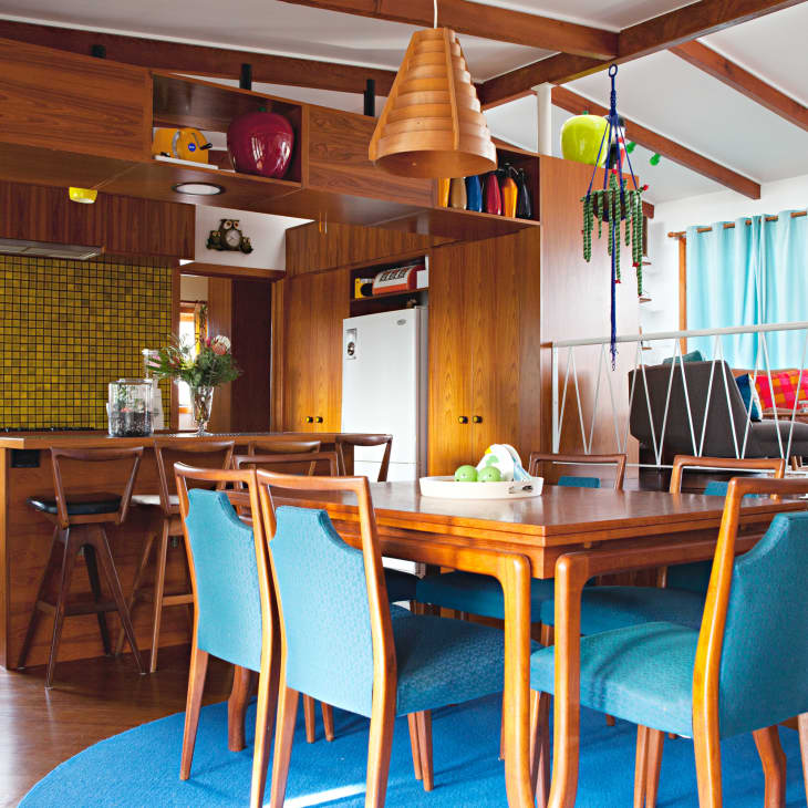 A mid-century kitchen/dining room full of rich brown wood furniture. The dining furniture features robin egg blue fabric and a circular rug underneath matches