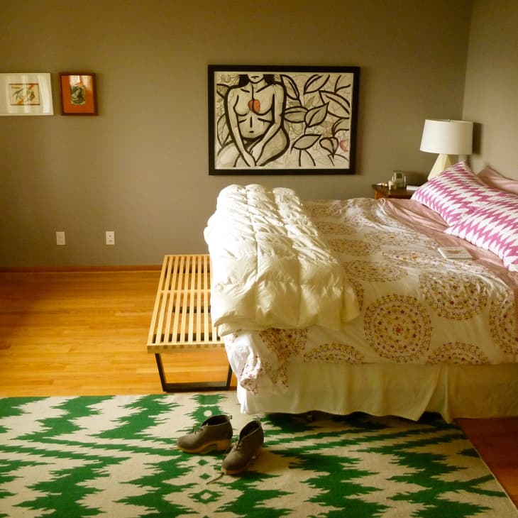 Bedroom with a patterned bedspread, pink pillows, abstract wall art, wooden bench, and green geometric rug.