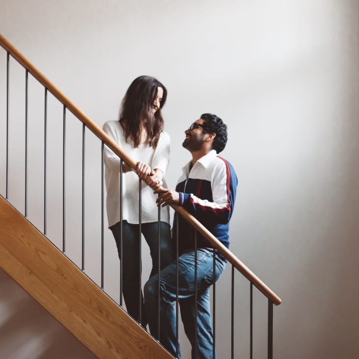 A couple standing on a wooden staircase, smiling at each other, with exposed brick walls.