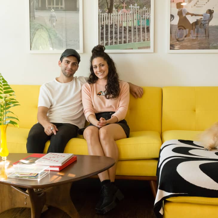 Couple sitting on a yellow sofa with a small dog, wooden coffee table, and framed art on the wall.