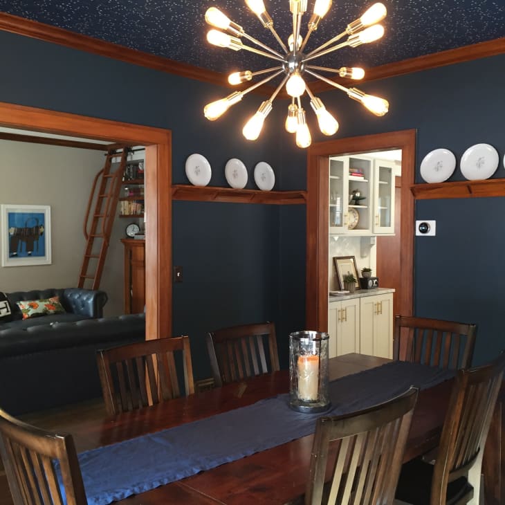 Dining room with dark wood table, modern chandelier, navy walls, and decorative plates on a shelf.