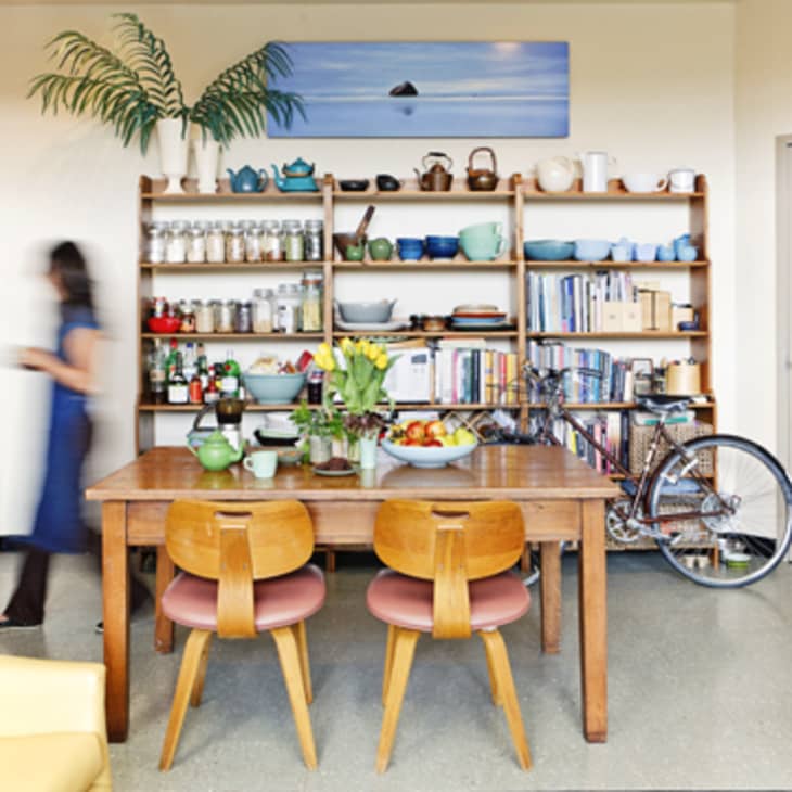 Cozy kitchen with wooden table, colorful chairs, open shelves, and a bicycle. Woman in blue dress walking by.