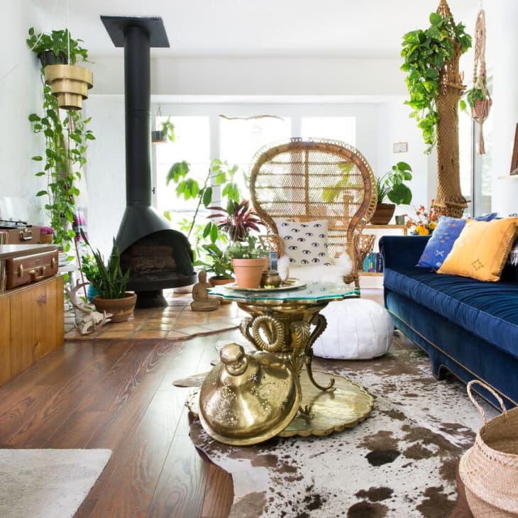 Eclectic living room with blue velvet sofa, peacock chair, gold table, wood stove, and lush plants.