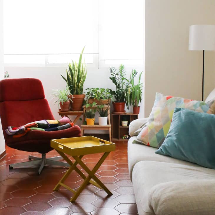 Living room with red swivel chair, beige sofa, colorful pillows, yellow tray table, and potted plants by the window.