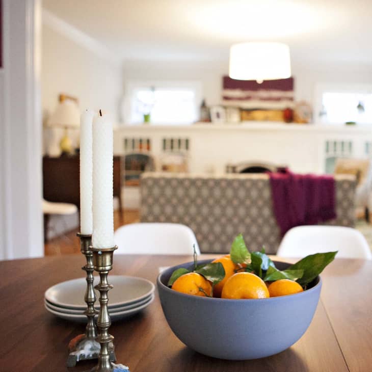 Dining table with a bowl of oranges and candlesticks, overlooking a cozy living room with a patterned sofa and purple throw.