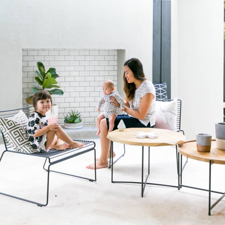 Woman with baby and child sitting on patio chairs, surrounded by plants and modern tables with decorative pots.