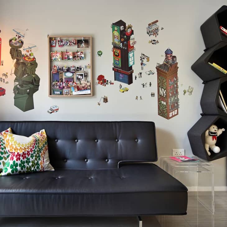 Black leather sofa with colorful pillows, wall art of cityscapes, hexagonal shelves, and a clear side table with a toy.