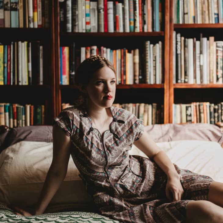 Woman in patterned dress sitting on a bed, surrounded by bookshelves filled with various books.