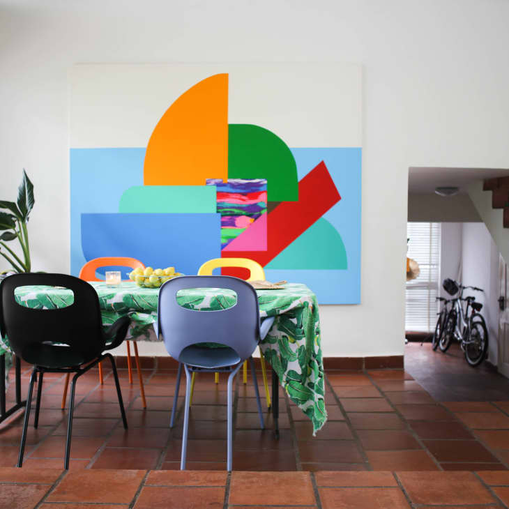Dining room with colorful abstract art, green tablecloth, black and blue chairs, and a plant by large windows.