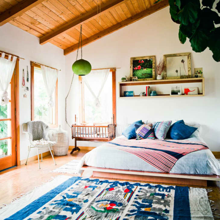 Cozy bedroom with wooden ceiling, colorful bedding, hanging crib, and decorative shelf above the bed.