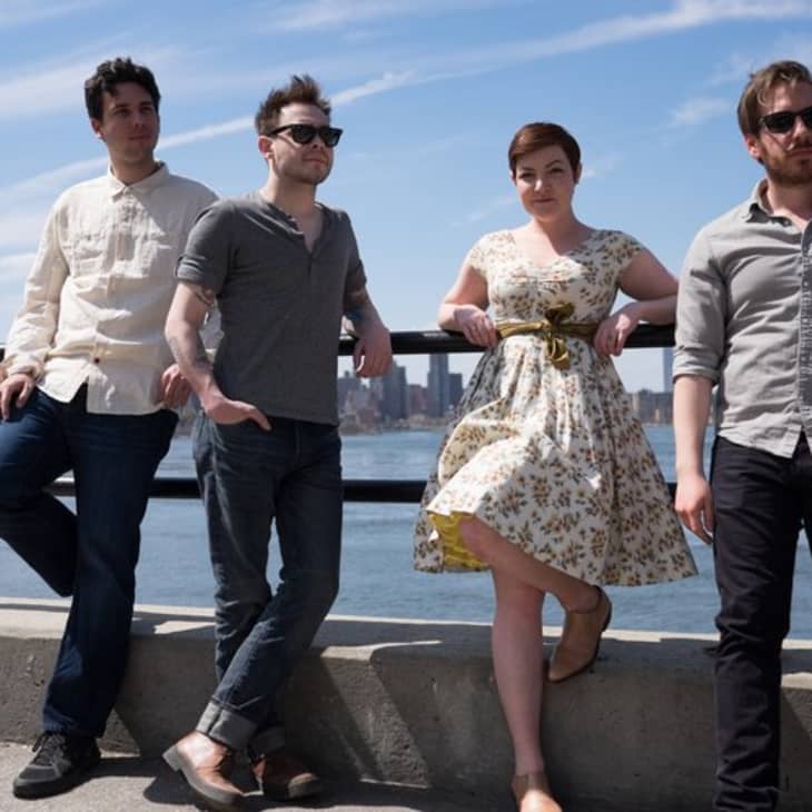 Four people standing by a waterfront railing, city skyline in the background.