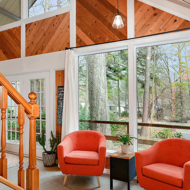 Living room with large windows, two orange armchairs, a small table, and potted plants, overlooking a wooded area.