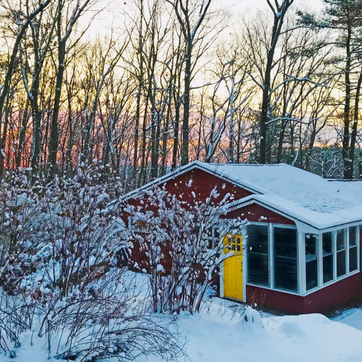 Red cabin with a yellow door surrounded by snow-covered trees at sunset.