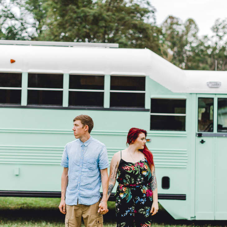 Couple holding hands in front of a mint green converted school bus with trees in the background.