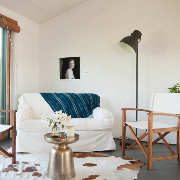 Cozy living room with white sofa, blue throw, wooden chairs, floor lamp, and a small round table with flowers.