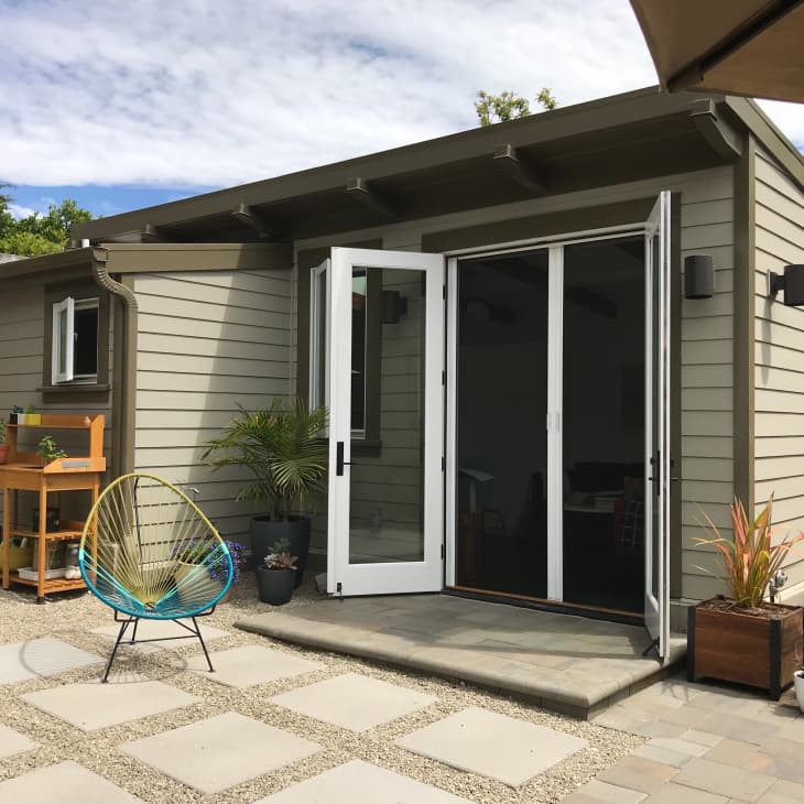 Small backyard studio with open French doors, two colorful chairs, potted plants, and a wooden potting bench.