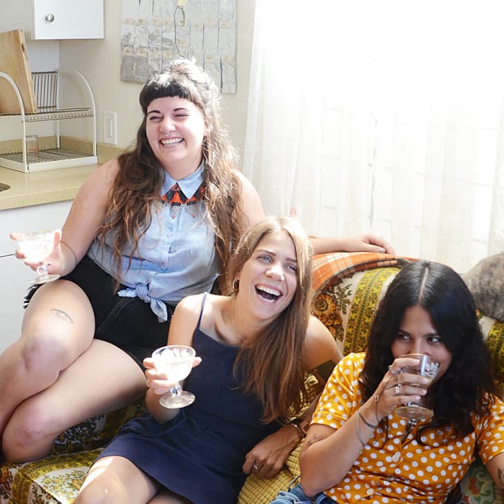 Three women laughing and holding drinks on a patterned sofa, with a cat lounging beside them in a cozy kitchen.