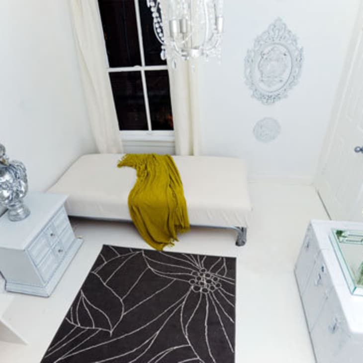 Minimalist white room with a single bed, yellow throw, black floral rug, and decorative silver bust.