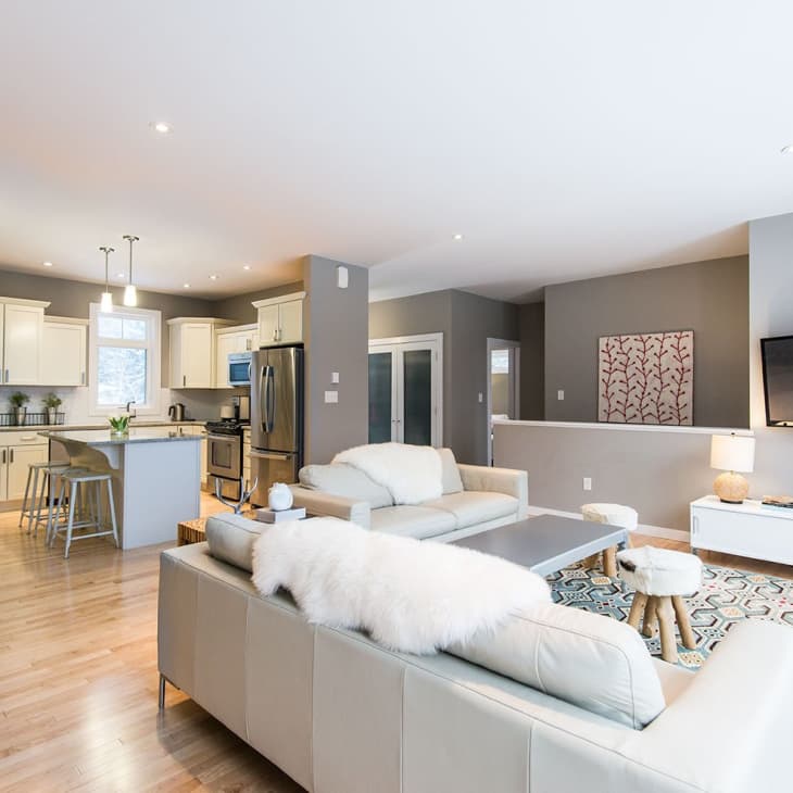 Open-concept living room and kitchen with beige sofas, white cabinets, stainless steel appliances, and a wall-mounted TV.