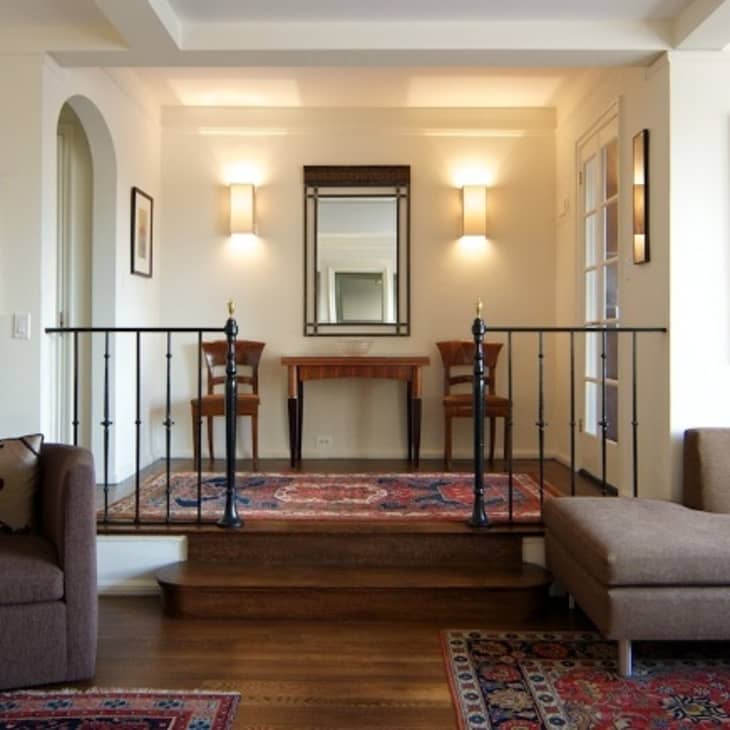Living room with brown sofas, ornate rugs, a console table, and a mirror flanked by two chairs under warm wall lights.