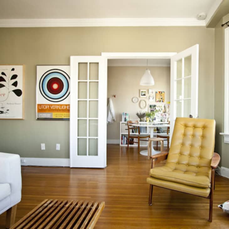 Living room with a mustard armchair, white sofa, and art on walls, leading to a dining area through French doors.